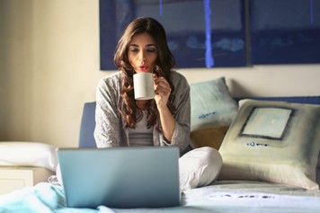 A woman in a grey jacket sits on the bed and uses a grey laptop.