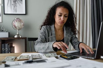 A woman in a plaid blazer using her laptop and calculator to count money.